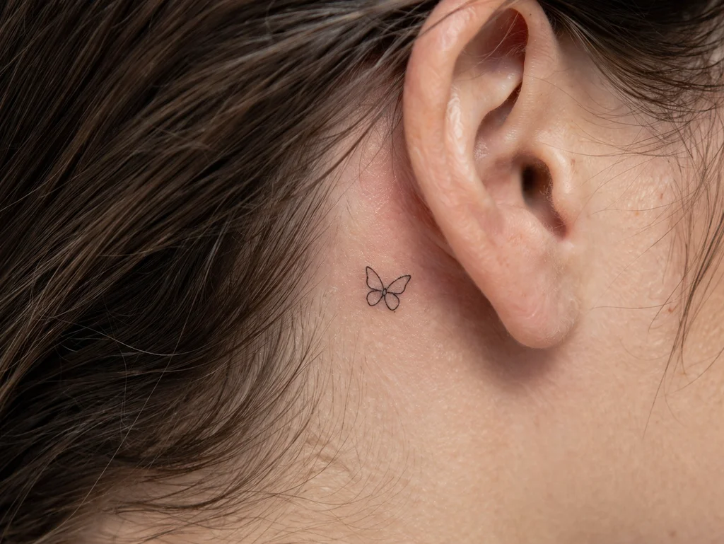 A tiny minimalist butterfly outline tattoo behind a womans ear in delicate thin black lines