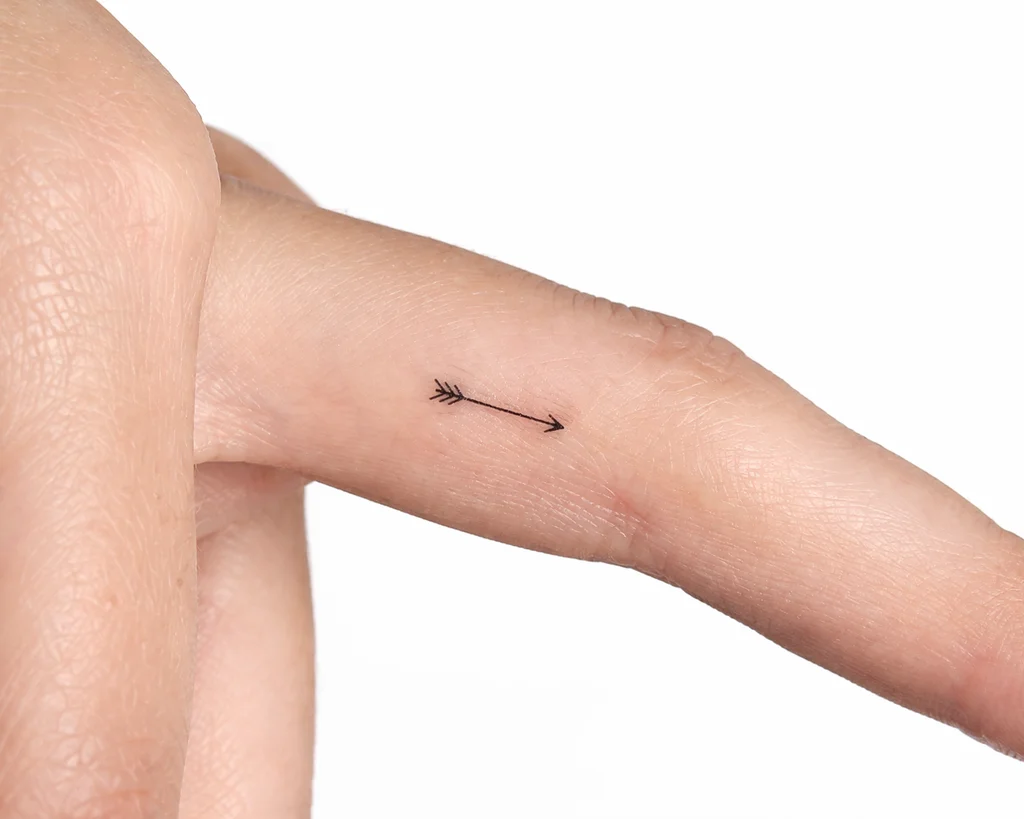A tiny minimalist single-line arrow tattoo on the side of a finger in clean black ink