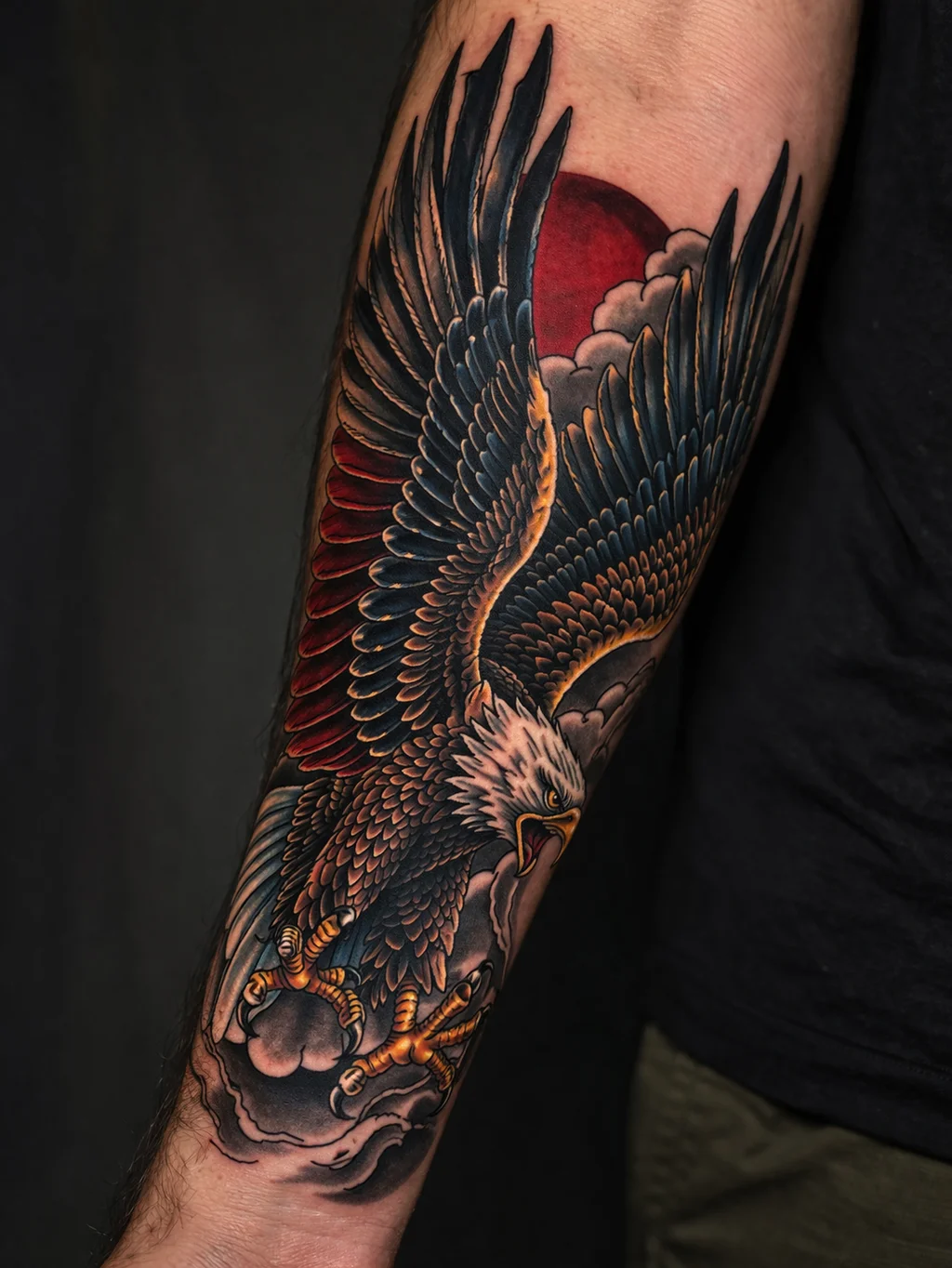 A neo-traditional eagle tattoo in flight on a mans outer forearm with bold outlines and vivid red blue and gold colors