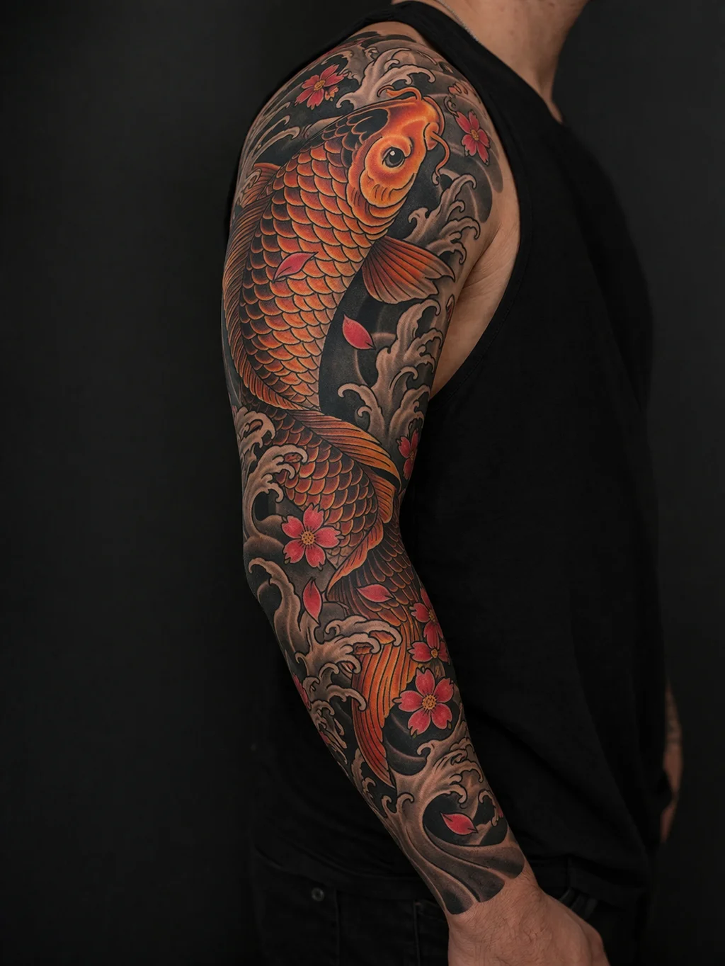 A traditional Japanese koi fish tattoo on a mans full arm sleeve with orange and gold fish swimming through waves and cherry blossom petals in Irezumi style